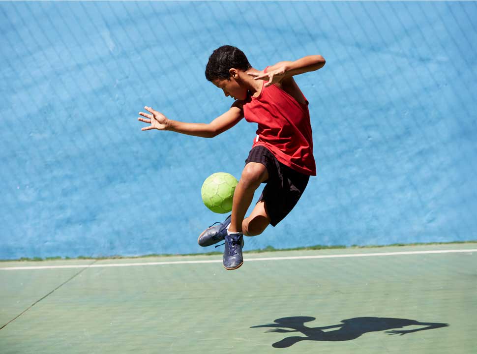 A young boy in a red shirt leaps in the air, kicking a ball