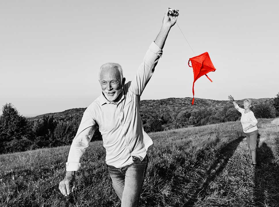 Behavioral-Science-Biometric-Research A smiling older man runs through a grassy hillside field flying a red kite while an older woman reaches up toward it, in a black-and-white photo with the kite highlighted in red.