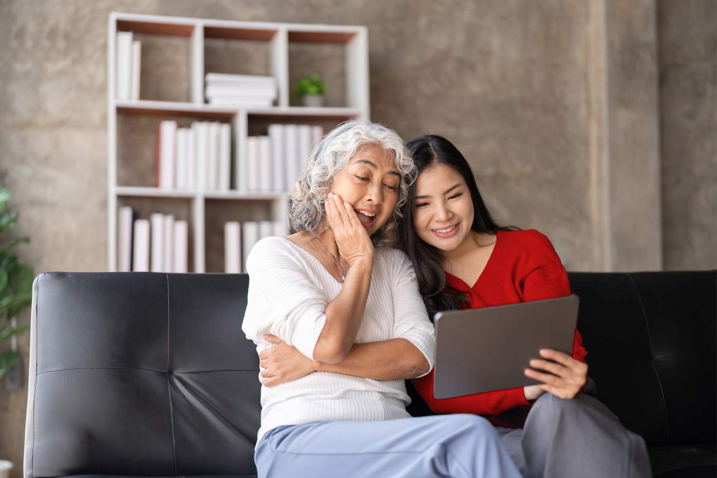 A mother and adult daughter of Asian descent sit on a couch and look at a tablet device together.