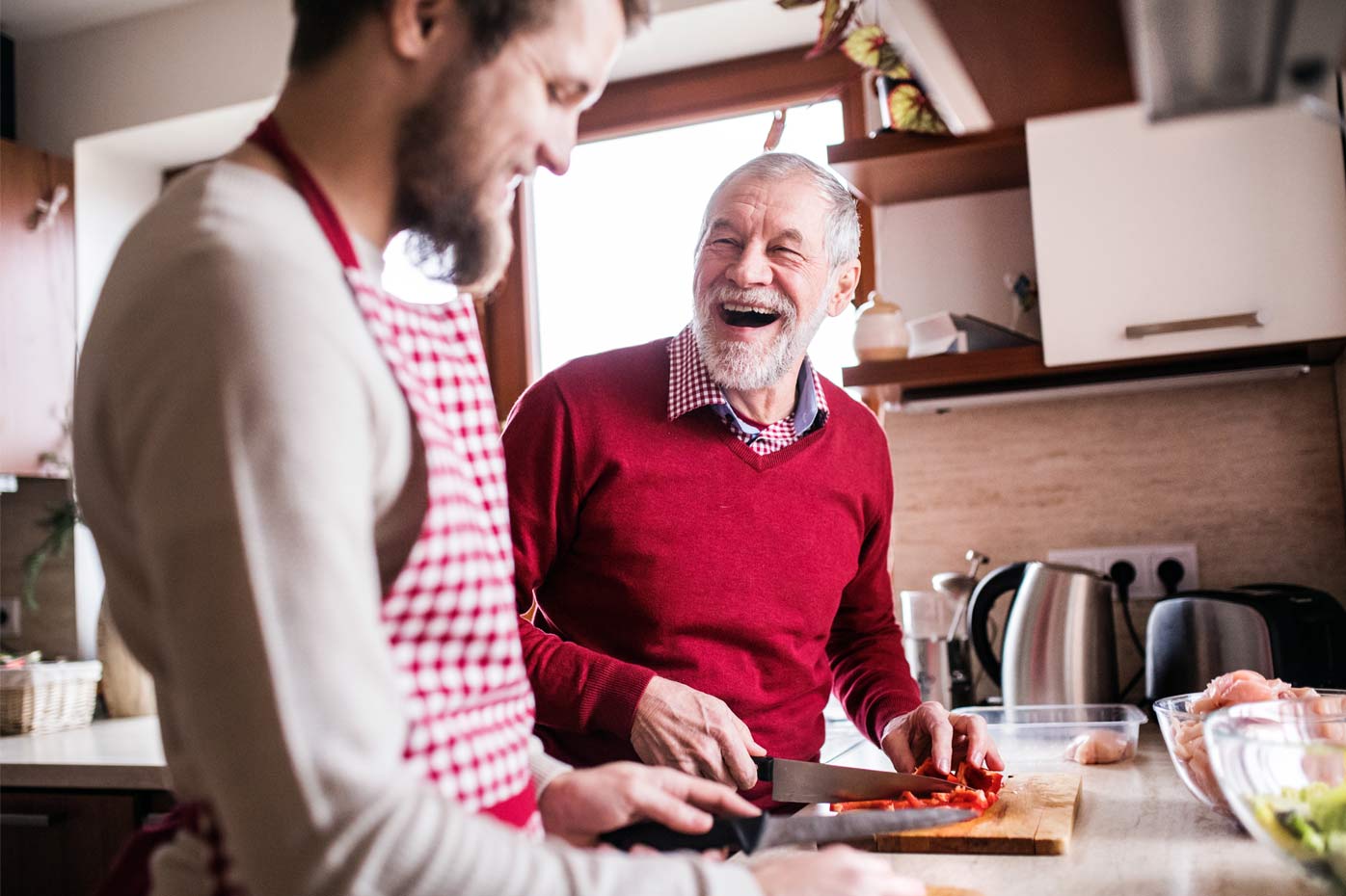 An older father in a red sweater cooks in the kitchen with his son, who wears an apron.