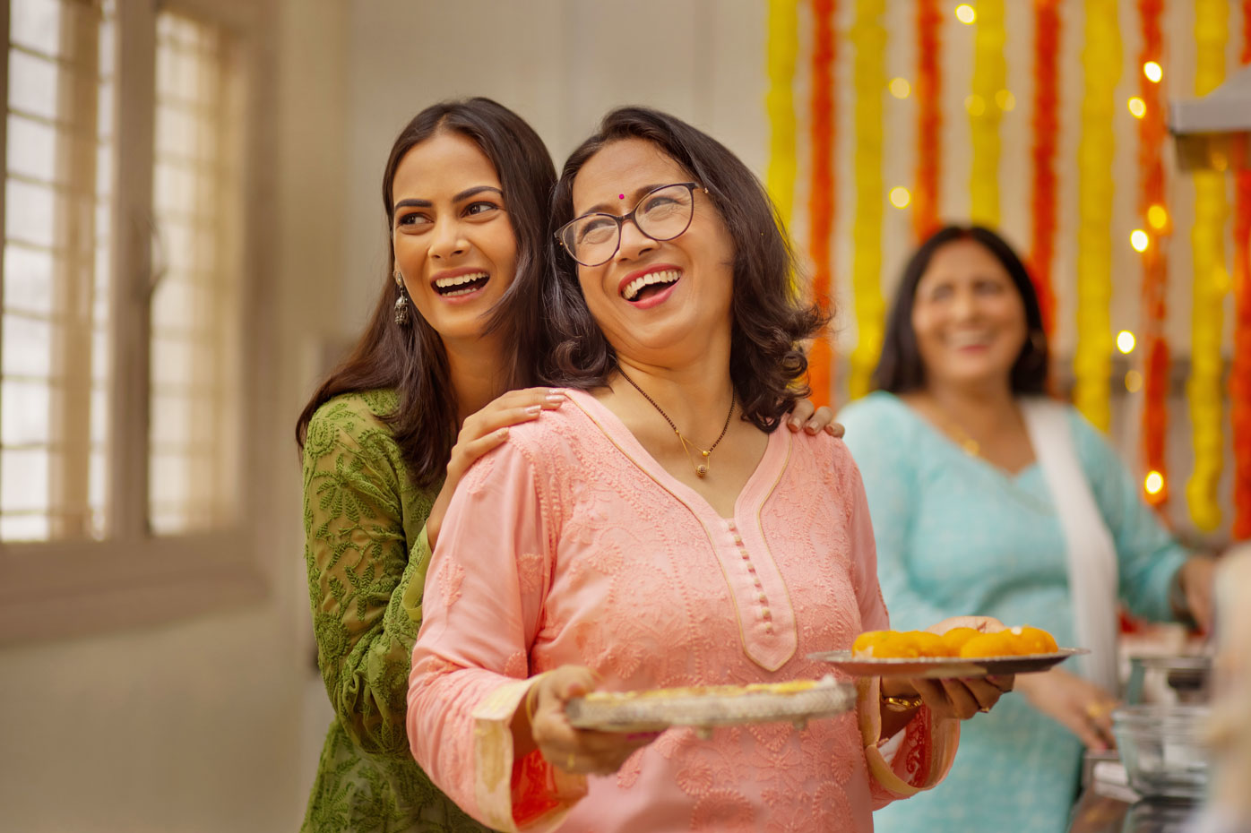 A smiling Indian mother is embraced from behind by her grown daughter.