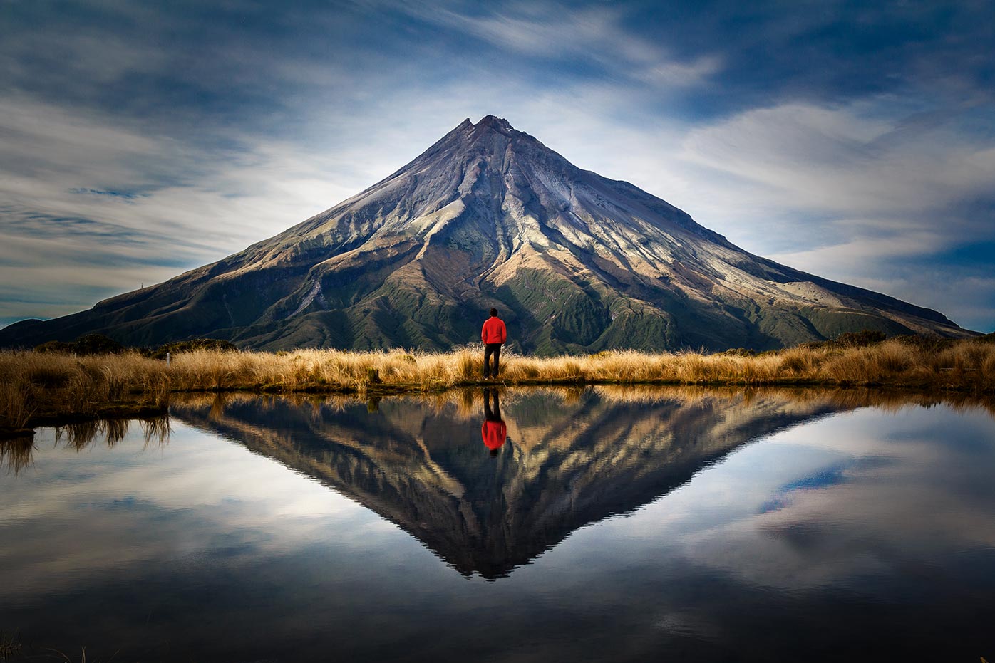 A person standing in front of a mountain with their reflection showing in nearby water